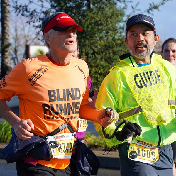 Visually impaired man running next to a sighted guide, joined by a running tether.